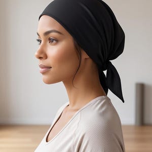 Könnte beinhalten: Ein einfarbig schwarzes Kopftuch, das im Nacken mit zwei kurzen Stoffbändern gebunden ist. Die Kopfbedeckung besteht aus einem glatten, blickdichten Material, das eng am Kopf anliegt und das Haar vollständig bedeckt, wobei die Ohren teilweise sichtbar bleiben.