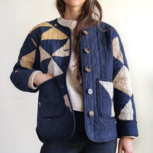 May include: A quilted jacket in navy blue, beige, and tan. The jacket features a geometric pattern on the shoulders and sleeves, with a solid navy blue body. It has large pockets and wooden buttons. The jacket is a stylish outerwear option.