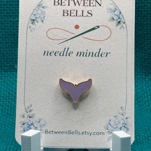 May include: A lavender whale tail needle minder with a gold-toned metal edge, mounted on a white display card featuring the text BETWEEN BELLS needle minder and BetweenBells.etsy.com, decorated with blue floral illustrations at the corners.