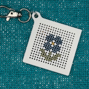 May include: A white, square cross-stitch keychain featuring a blue and yellow flower design with green leaves. The keychain has a silver clasp and ring for attaching to keys or bags. The background is a teal fabric.