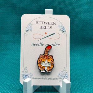 May include: An orange tabby cat enamel needle minder pin featuring a grumpy expression and a raised tail. The pin is attached to a white display card printed with the text BETWEEN BELLS, needle minder, and BetweenBells.etsy.com with floral accents.