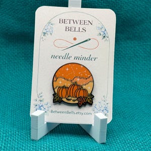 May include: A circular enamel needle minder featuring an orange sunset sky with white stars and clouds above three orange pumpkins and autumn leaves. The pin is attached to a white display card with the text BETWEEN BELLS and needle minder.