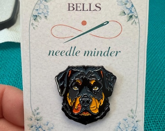 May include: A black and tan Rottweiler needle minder pin on a decorative card. The pin features a detailed dog head design. The card has the text "BETWEEN BELLS" and "needle minder" with a floral design. The Etsy shop URL is also visible.