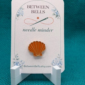 May include: A decorative needle minder with an orange seashell design. The card reads "Between Bells" and "needle minder" with a needle and thread graphic. The Etsy shop URL is visible at the bottom. The card is displayed on a white stand.