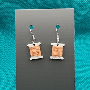 May include: A pair of earrings shaped like miniature thread spools. Each earring has a white plastic spool shape wrapped with orange thread, hanging from a silver-coloured fish hook. The earrings are displayed on a dark grey card against a teal background.
