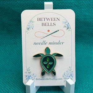 May include: A teal and gold turtle needle minder on a white card with the text "BETWEEN BELLS" and "needle minder". The card is displayed on a white easel. The Etsy shop URL is visible on the card.