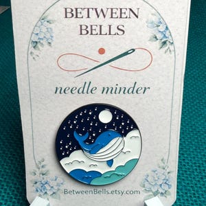 May include: A circular needle minder featuring a blue whale design. The whale is set against a dark blue night sky with white clouds and a full moon. The card reads "BETWEEN BELLS" and "needle minder".