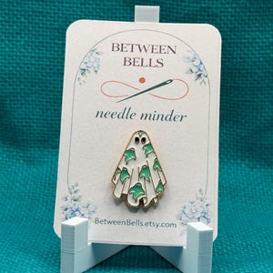 May include: A white ghost-shaped enamel needle minder featuring a pattern of small green ghosts, displayed on a white card labeled BETWEEN BELLS needle minder with the website BetweenBells.etsy.com printed at the bottom of the card.