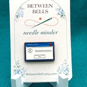 Puede incluir: Un sujetador de agujas con un diseño de mensaje de error de Windows XP. El pin dice "Tarea fallida con éxito" con un botón "OK". La tarjeta dice "Between Bells" y "needle minder" con un diseño floral.