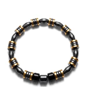 Puede incluir: Una pulsera de cuentas con cuentas grises oscuras y negras alternas con detalles dorados. La pulsera está hecha de múltiples cuentas redondas y cilíndricas, creando un accesorio elegante. La pulsera se muestra sobre un fondo blanco.