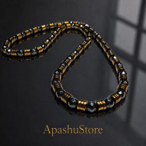 May include: A necklace featuring alternating dark grey faceted beads and cylindrical gold-toned metal spacers. The jewellery piece is arranged in a circular loop, showcasing a repeating pattern of metallic and stone-like elements. Text reads ApashuStore.