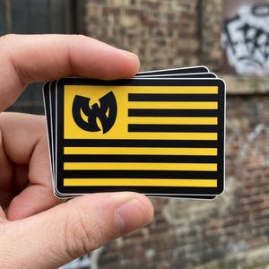 May include: A stack of rectangular stickers featuring a yellow and black flag design. The flag has horizontal black stripes and a yellow emblem in the upper left corner. The stickers are held in a hand.