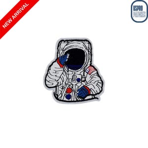 May include: Embroidered patch featuring an astronaut saluting. The patch is white with black details, red and blue accents, and an American flag on the shoulder. The words "NEW ARRIVAL" are in red on a diagonal banner.
