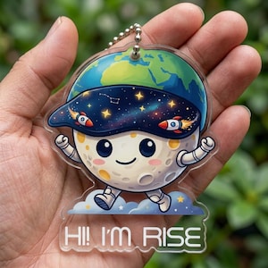 May include: A clear acrylic keychain featuring a cartoon moon character wearing a space-themed visor and a smiling face. The visor has rockets and stars. The keychain has the text "HI! I'M RISE" at the bottom. The moon is walking on a cloud.