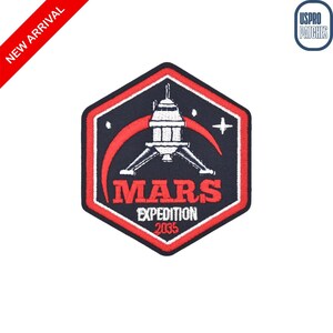 May include: A hexagonal patch with a red and black design. The patch features a spacecraft illustration, the word "MARS" in red, and the text "EXPEDITION 2035". A red banner reads "NEW ARRIVAL".