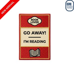 May include: A rectangular patch with a red background and gold trim. The patch features the text "BOOK LOVER" in a white oval at the top, and "GO AWAY! I'M READING" in black text. An open book graphic is at the bottom.