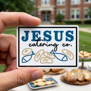 May include: A rectangular sticker with the text "JESUS catering co." in blue and black. The sticker features illustrations of bread loaves and fish in blue and tan. The sticker is held in a person's hand.
