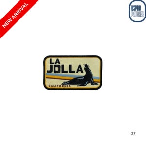 May include: Rectangular patch featuring a black sea lion silhouette, the words "LA JOLLA" in bold black letters, and "CALIFORNIA" in a smaller font. The background is beige with horizontal stripes of blue, yellow, and brown.