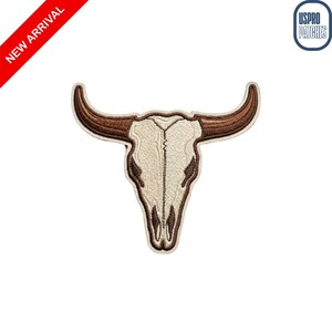 May include: An embroidered patch featuring a steer skull design with a cream-colored cranium and dark brown horns. The patch has a distinct stitched border and is presented against a plain white background with a red corner banner and logo.