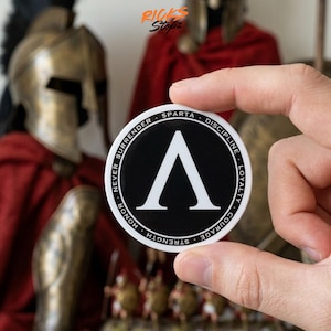 May include: A round, black and white sticker featuring a white Lambda symbol and the words "Sparta," "Discipline," "Loyalty," "Courage," "Strength," "Honor," and "Never Surrender." The sticker is held in a hand, with a blurred background of Spartan helmets and red cloaks.