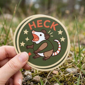 May include: A round sticker featuring a cartoon opossum with an open mouth, wearing a green shirt. The sticker has the word "HECK" in orange letters and is surrounded by stars. The opossum has a brown body, white face, and a striped tail.