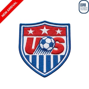 May include: A shield-shaped embroidered patch featuring a red top section with three white stars, and a white middle section with the red letters U and S flanking a central soccer ball. The bottom section has vertical blue and white stripes.