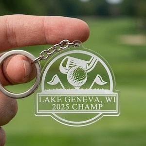 Puede incluir: Llavero de golf de acr&iacute;lico transparente con un llavero y cadena plateados. El llavero presenta un dise&ntilde;o de pelota de golf, palo y banderas, con el texto "LAKE GENEVA, WI 2025 CHAMP". El fondo es verde borroso.