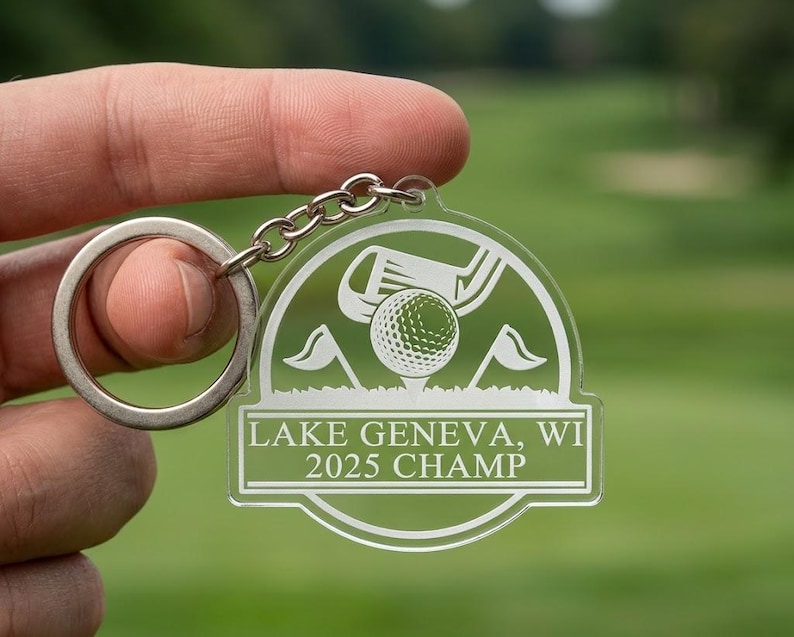 Puede incluir: Llavero de golf de acr&iacute;lico transparente con un llavero y cadena plateados. El llavero presenta un dise&ntilde;o de pelota de golf, palo y banderas, con el texto "LAKE GENEVA, WI 2025 CHAMP". El fondo es verde borroso.