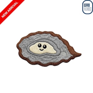 May include: Embroidered patch featuring a smiling oyster design with a light beige interior and a gray and brown exterior. The patch has a cartoon-like appearance and is labeled "NEW ARRIVAL". The patch is a fun accessory.