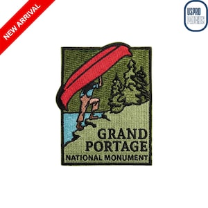 May include: Embroidered patch featuring a person carrying a red canoe over their head, with the text "GRAND PORTAGE NATIONAL MONUMENT." The patch has a green background with a blue river and trees.