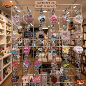 Puede incluir: Una exhibici&oacute;n de llaveros del D&iacute;a de la Madre con varios dise&ntilde;os y frases como "Mother", "Love Mama" y "Best Mom". Los llaveros est&aacute;n en una rejilla de metal, con un letrero de madera que dice "Handmade Mama Gifts Mother's Day Keychains."