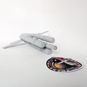 May include: A light gray model of a spacecraft with multiple cylindrical engines and wings. Next to it is a colorful sticker with the text "PROJECT HAIL MARY" and a design featuring a rocket and flags.