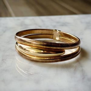 May include: A gold-toned bracelet with a flexible, ribbed design. The bracelet wraps around the wrist twice, with a smooth, oval-shaped accent. The jewelry is displayed on a white marble surface.