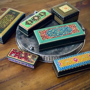May include: A collection of decorative boxes with intricate floral and geometric designs. The boxes are in various shapes and sizes, featuring colors like green, red, blue, and gold. They are arranged on a metal tray and a wooden surface.