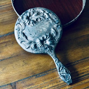 May include: An antique silver-toned hand mirror with an ornate floral design around the circular mirror and on the handle. The mirror is resting on a wooden surface, with a decorative tray in the background.