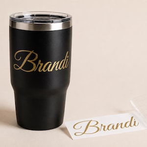 May include: A matte black stainless steel tumbler with a clear lid, featuring the name Brandi in gold cursive script. Beside the tumbler lies a matching gold vinyl decal of the name Brandi on a white backing, ready for application to a surface.