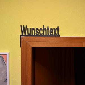 Könnte beinhalten: Ein schwarzes, lasergeschnittenes Schild mit dem deutschen Wort "Wunschtext" in einer serifenlosen Schriftart. Das Schild ist an der oberen Ecke eines hölzernen Türrahmens vor einer hellgelben Wand befestigt und ragt leicht über den Rahmen hinaus.