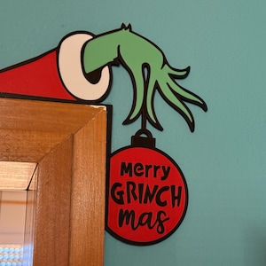 Könnte beinhalten: Ein dekoratives Wandschild mit einer grünen Hand, die aus einer rot-weißen Weihnachtsmütze ragt und eine runde rote Weihnachtskugel hält. Die Kugel trägt den schwarzen Schriftzug merry GRINCH mas. Das Design hat durchgehend schwarze Konturen.