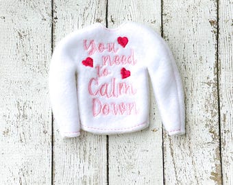 以下が含まれることがあります： ピンク色の刺繍で「You need to Calm Down」と書かれた、3つの赤いハートが付いた白いフェルトセーター。セーターはクルーネックと長袖です。