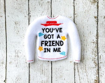 以下が含まれることがあります： 「YOU'VE GOT A FRIEND IN ME」の黒い文字が入った、白いフェルトのセーター型オーナメント。赤い帽子、青い雲、黄色の星のデザイン。縁は赤いステッチでトリミングされています。
