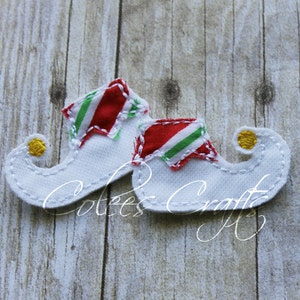 May include: A pair of white felt elf shoes with red and green striped accents on the cuffs. The shoes have a curled toe with a yellow pom-pom and are stitched with white thread. The design is festive and suitable for holiday crafts.