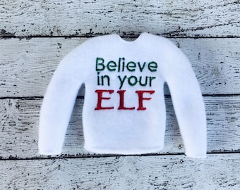 以下が含まれることがあります： 白いフェルトのセーター、長袖。 「Believe in your ELF」の文字が緑と赤で刺繍されています。 このセーターはホリデーテーマ用にデザインされています。