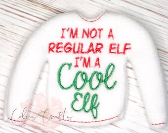 以下が含まれることがあります： 「I'M NOT A REGULAR ELF I'M A Cool Elf」と赤と緑の刺繍が施された白いフェルトセーター。セーターは赤いステッチの輪郭と袖が付いています。