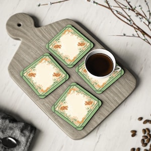 Könnte beinhalten: Vier quadratische Untersetzer mit grünem Rand und cremefarbenem Mittelteil, jeweils mit einem dekorativen Design mit orangefarbenen und grünen Akzenten. Eine weiße Kaffeetasse mit dunkler Flüssigkeit steht auf einem Untersetzer, platziert auf einem Holzschneidebrett.