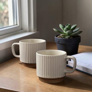 May include: Two cream-coloured ceramic mugs with vertical ribbed detailing and a brown base. The mugs have a handle and are placed on a wooden surface. A small succulent plant in a black pot is in the background.
