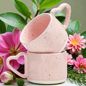 Puede incluir: Dos tazas de cerámica rosa apiladas, cada una con un asa en forma de corazón. Las tazas tienen un diseño moteado y se colocan frente a flores rosas y follaje verde. Las tazas tienen una base blanca.