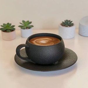 Può includere: Una tazza da caffè e un piattino in ceramica neri. La tazza è piena di caffè con un disegno bianco. La tazza ha una superficie strutturata e una forma arrotondata. Il piattino è rotondo e ha una trama abbinata.