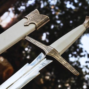 May include: A silver-coloured metal sword with a white grip and a matching white scabbard. The sword features an ornate, gold-toned metal crossguard and pommel with intricate engraved patterns. The scabbard includes a decorative gold-toned metal throat.