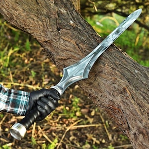 May include: A double-edged sword with a polished silver-coloured metal blade featuring a central ridge and a flared guard. The handle is wrapped in black material with a rounded silver-coloured pommel at the base, held by a gloved hand.