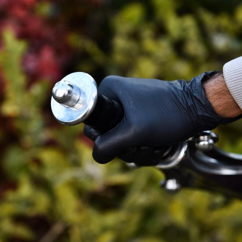 May include: Close-up of a black handlebar grip with a silver cap. The grip is held by a hand wearing a black glove. The background is blurred with green and red foliage.